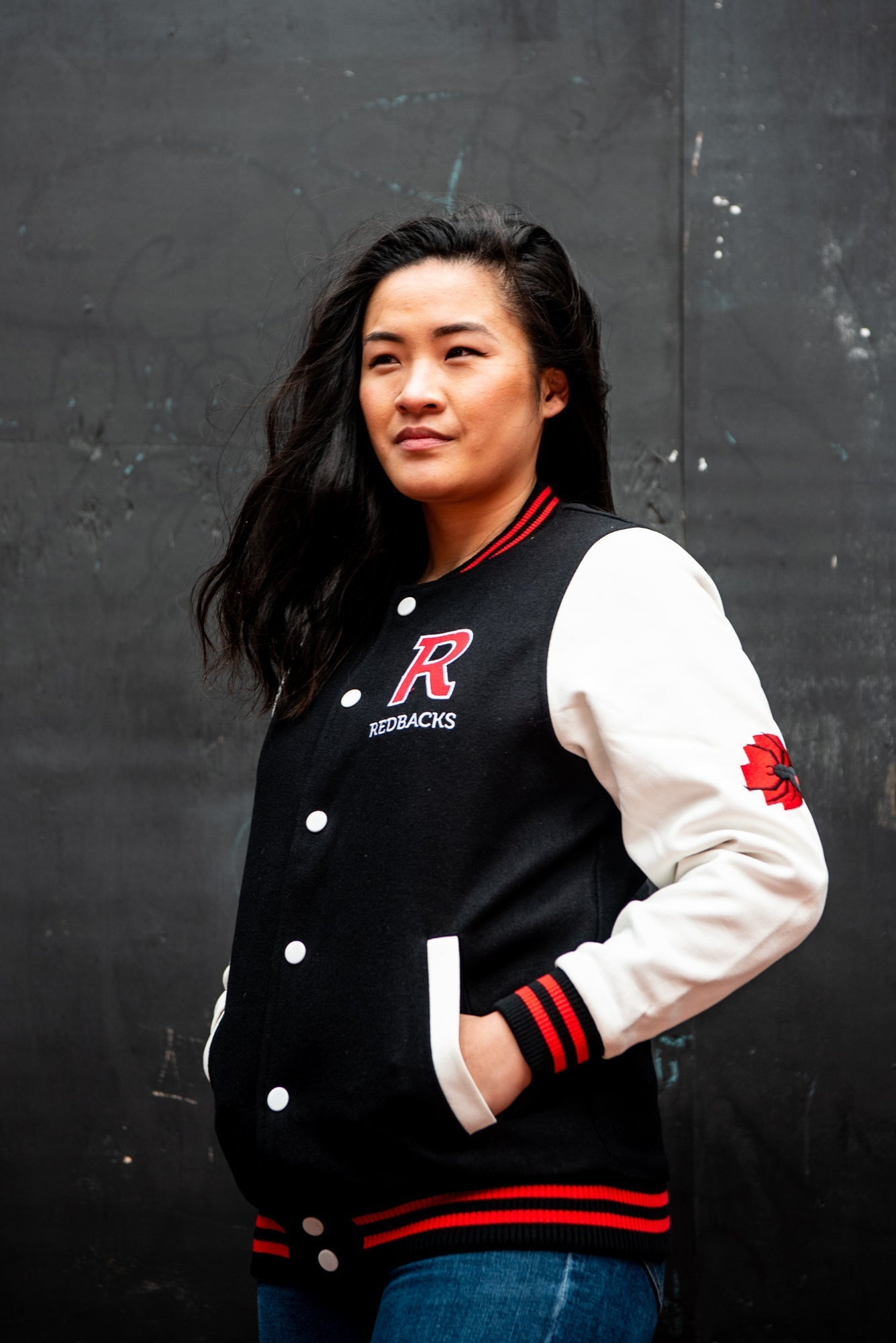 Female model wears an RMIT Store black varsity jacket with white sleeves, black collar and hem featuring red stripe detailing, and a large red chenille letter "R" patch on the left chest; white snap buttons fasten the front, and the model is posed in a front angled view looking to her right.
